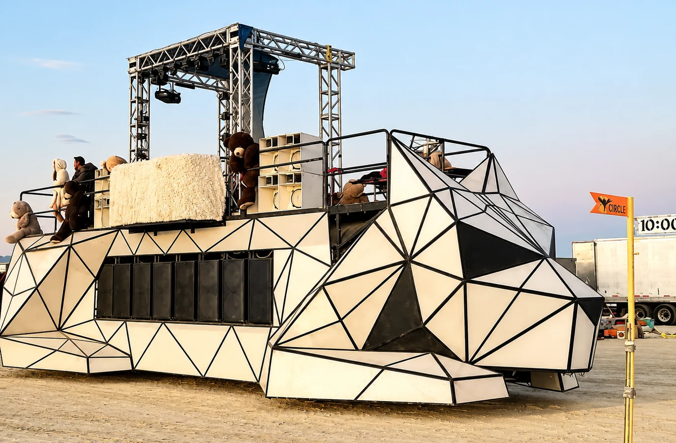 The Dogzilla art car on the Black Rock City playa: a geometric white and black body, a speaker wall, a truss stage on top with plush bears along the rail