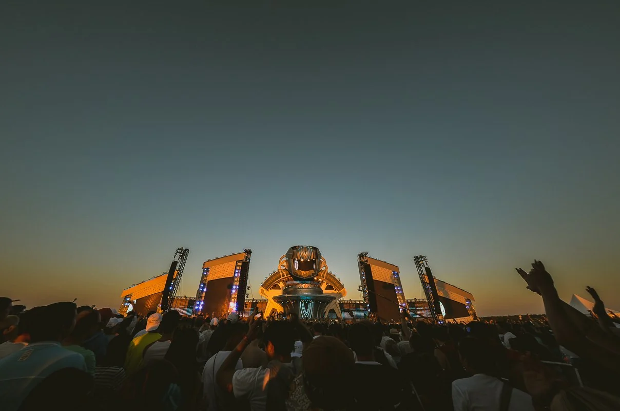 A festival main stage with LED towers and a sculpted centrepiece at sunset, a crowd in front with hands raised