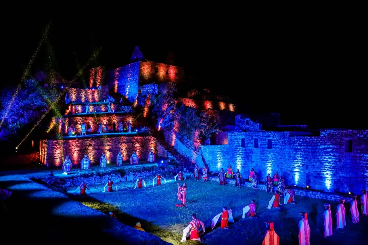 Performers in colourful costumes on a lawn below the stone terraces of Machu Picchu, lit blue and amber at night