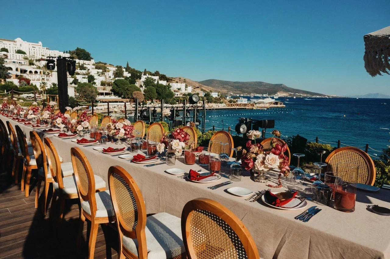 A long dinner table on a terrace over the sea, rattan chairs, red and white roses, burgundy napkins, speakers and lights rigged along the rail, white villas on the hillside behind
